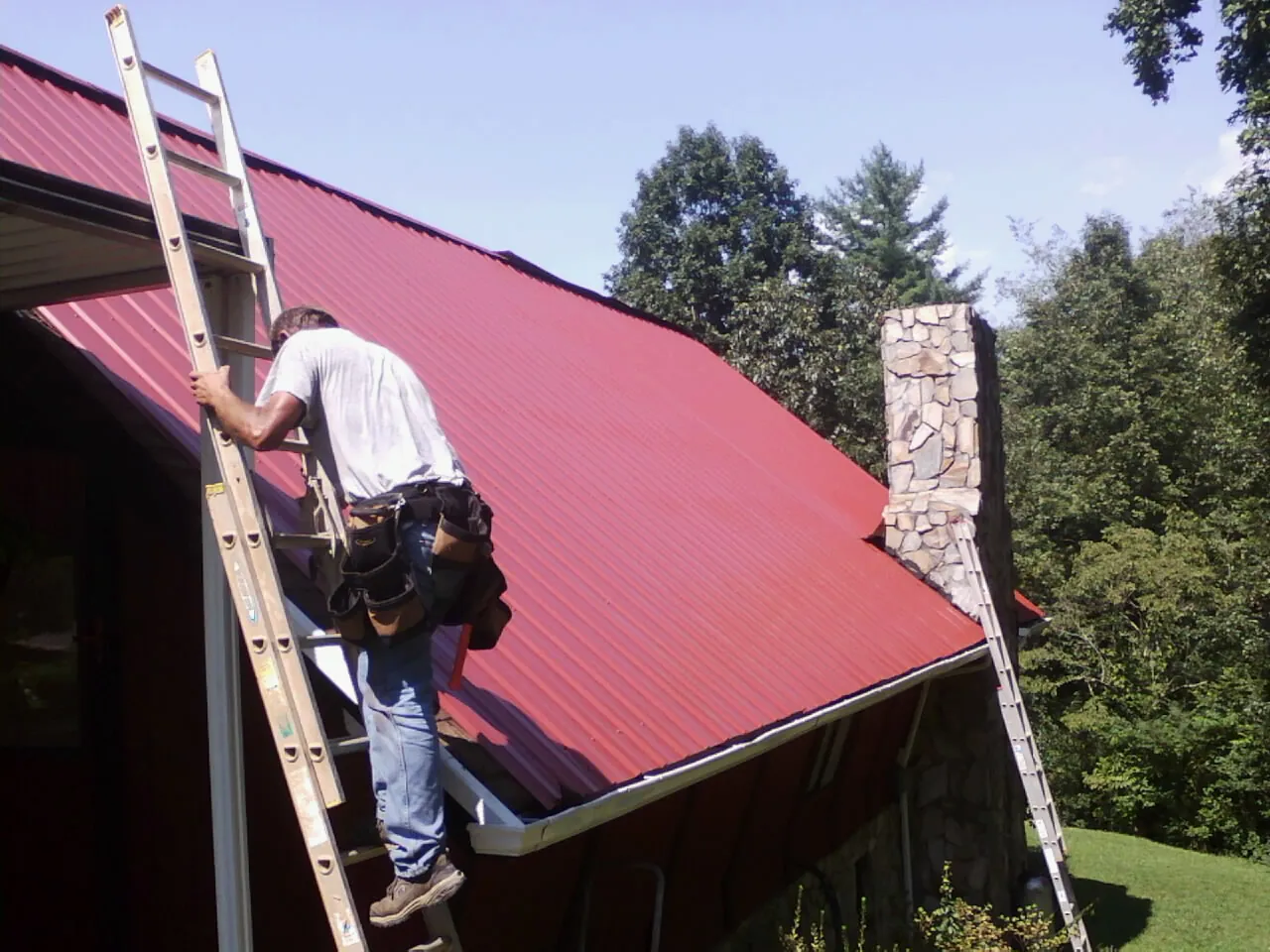 Roofer on a ladder beside a red metal roof and stone chimney in Anahuac